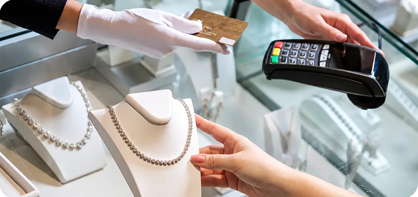 Jewelry store employee processing a customer payment with a POS terminal beside a diamond necklace display.